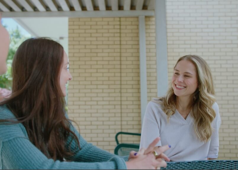 Two female students sitting on a picnic table smiling at eachother